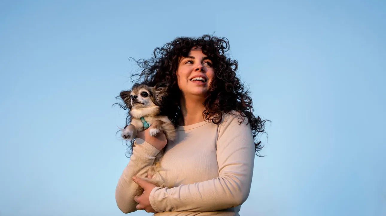 Mujer con cabello rizado sosteniendo a su perro chihuahua bajo un cielo azul."