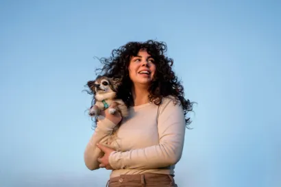 Mujer con cabello rizado sosteniendo a su perro chihuahua bajo un cielo azul."