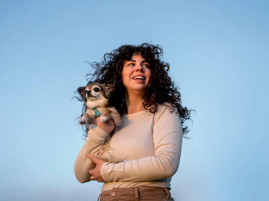 Mujer con cabello rizado sosteniendo a su perro chihuahua bajo un cielo azul."
